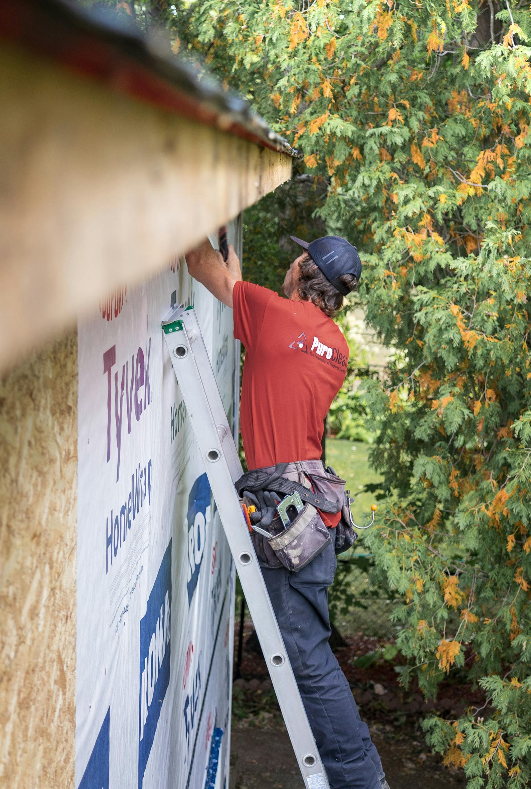 Adult male construction worker wearing a red shirt repairs roof siding outdoors with green trees in background.
